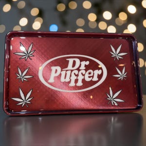 May include: A rectangular, red metal rolling tray with a metallic finish. The tray features the text "Dr. Puffer" in a white oval, surrounded by white cannabis leaf designs. The tray is designed for rolling herbs.