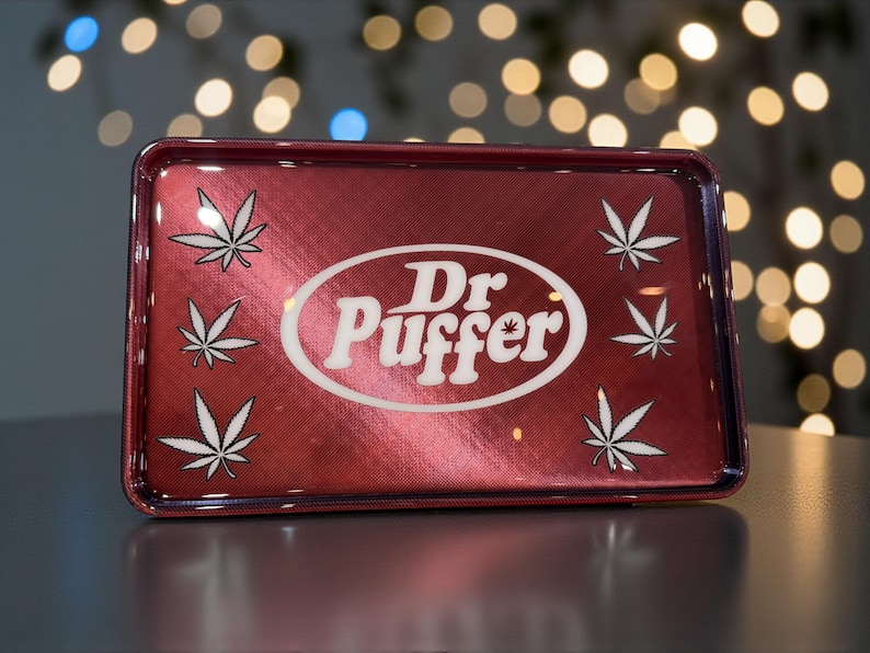May include: A rectangular, red metal rolling tray with a metallic finish. The tray features the text "Dr. Puffer" in a white oval, surrounded by white cannabis leaf designs. The tray is designed for rolling herbs.