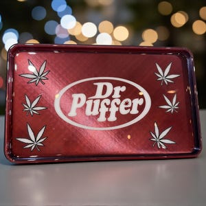 May include: A rectangular, red metal rolling tray with the text "Dr. Puffer" in a white oval, and white cannabis leaf designs. The tray has a metallic finish and a raised edge.