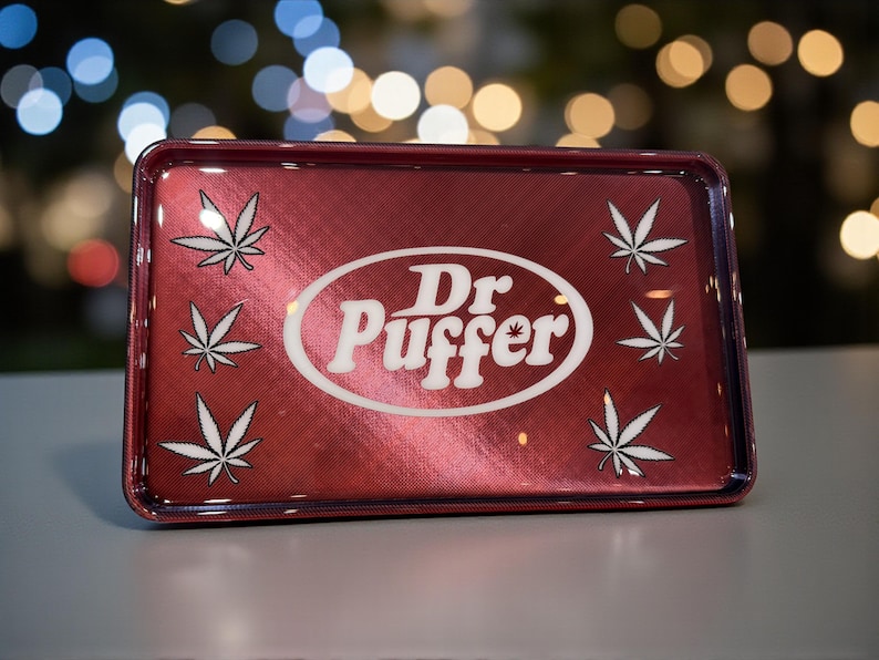 May include: A rectangular, red metal rolling tray with the text "Dr. Puffer" in a white oval, and white cannabis leaf designs. The tray has a metallic finish and a raised edge.
