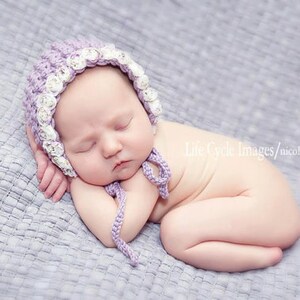 May include: A newborn baby wearing a lavender crocheted bonnet with white floral accents, sleeping peacefully on a gray blanket. The bonnet has ties under the chin. The baby is curled up, with arms and legs tucked in.
