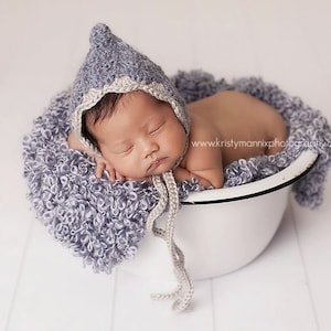 Può includere: Un neonato addormentato con un cappuccio di maglia grigio, appoggiato in una ciotola bianca foderata con una morbida coperta grigia e strutturata. Il cappuccio ha un bordo grigio chiaro e lacci.