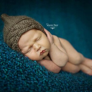 Può includere: Un neonato addormentato con un cappello marrone lavorato a maglia, adagiato su una coperta strutturata color turchese. Il bambino ha gli occhi chiusi e le braccia vicino al viso. Lo sfondo è di colore turchese uniforme.