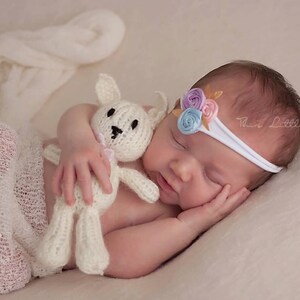May include: A sleeping baby wearing a white headband with pastel-colored fabric flowers, and a knitted white bunny toy. The baby is wrapped in a white blanket, and the image has a soft, neutral color palette.