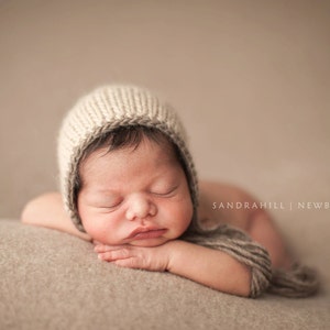 Puede incluir: Un bebé recién nacido durmiendo, con un gorro de punto beige claro, descansando sobre una manta suave de tono neutro. El bebé tiene los ojos cerrados y rasgos delicados. La imagen tiene un enfoque suave y una paleta de colores cálidos.