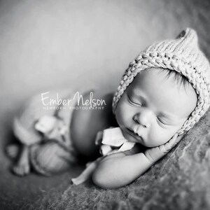 Puede incluir: Fotografía en blanco y negro de un recién nacido con gorro de punto, acostado sobre una superficie suave y texturizada. El bebé está acurrucado, con los ojos cerrados. La imagen incluye el texto "Ember Nelson Newborn Photography."