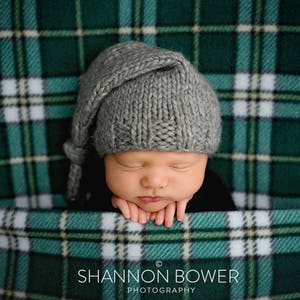 Op de afbeelding: Een pasgeboren baby met een grijze gebreide muts met een lange punt, rust vredig op een groen-witte geruite deken. De handen van de baby zijn zachtjes onder hun kin geplaatst. De deken heeft een donkergroen en wit ruitpatroon.