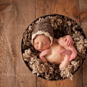 May include: A sleeping newborn baby wearing a brown and cream crocheted hat, lying in a round basket filled with soft, textured material. The baby is curled up. The background is a rustic wooden surface.