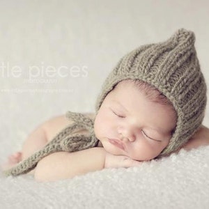 Puede incluir: Un recién nacido durmiendo con un gorro de punto verde oliva con una parte superior puntiaguda y una corbata a juego. El bebé está acostado sobre una superficie blanca y suave. La imagen tiene una estética suave y soñadora, ideal para fotografía de recién nacidos.