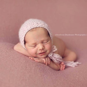 Op de afbeelding: Een pasgeboren baby met een lichtroze gebreide muts met een bijpassende strik, liggend op een zachte, roze deken. De baby glimlacht met gesloten ogen. De afbeelding is van cNewborn Daydreams Photography | 2014.