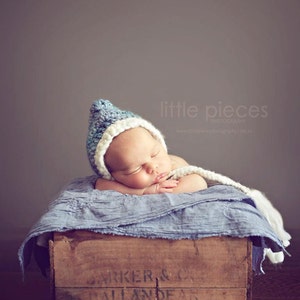May include: A sleeping baby wearing a blue and white knit cap and a white scarf, resting on a blue blanket inside a wooden crate. The crate has the text "BARKER & CO. BALLANDEAN" printed on it. The background is a solid gray.