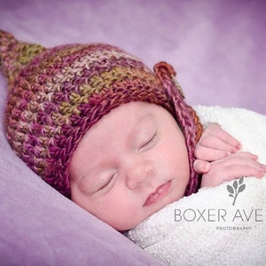 Könnte beinhalten: Ein schlafendes Baby, das eine handgefertigte, mehrfarbige Häkelmütze mit spitzer Spitze trägt. Das Baby ist in eine weiße Decke gehüllt und ruht auf einer lila Oberfläche. Das Bild enthält den Text "BOXER AVENUE PHOTOGRAPHY".