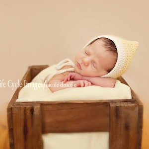 Peut inclure: Un nouveau-né endormi portant un bonnet en crochet jaune et blanc, dormant paisiblement dans une caisse en bois. Le bébé repose sur une couverture crème. La caisse est en bois brun foncé.