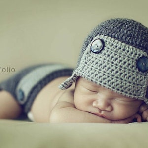 May include: A sleeping newborn baby wearing a handmade grey crochet hat with two large blue buttons and a matching nappy cover. The baby is lying on its front, eyes closed. The hat has ear flaps.