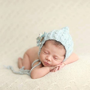 May include: A newborn baby wearing a pale blue knitted bonnet with a floral detail, lying on a cream blanket. The baby is curled up, resting its head on its hands.