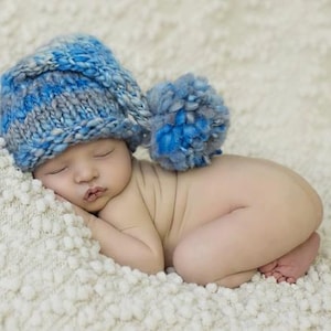 May include: A newborn baby asleep, wearing a blue and grey knitted hat with a large pom-pom. The baby is lying on a soft, white, textured blanket. The hat is made of blue and grey yarn.