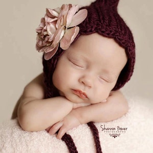 Può includere: Un bambino che dorme con un cappello di maglia bordeaux con un grande fiore rosa chiaro. Il bambino riposa su una superficie morbida rosa chiaro, con le braccia incrociate. Il cappello ha un cordino decorativo. L'immagine è una fotografia professionale.
