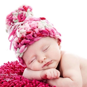 May include: A sleeping baby wearing a pink, white, and red crocheted hat with a ribbon. The baby is resting on a pink textured blanket. The image is set against a white background.