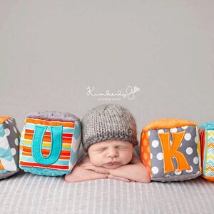 Puede incluir: Un bebé recién nacido con un gorro de punto gris descansa entre cinco bloques de tela coloridos que deletrean el nombre "LUKE". Los bloques presentan varios patrones, incluyendo chevrones, lunares y rayas, con acentos naranjas y turquesas.