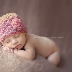 May include: A sleeping baby wearing a pink and cream crocheted hat with a flower detail. The baby is lying on a soft, textured surface. The image has a soft, diffused lighting and a neutral background.
