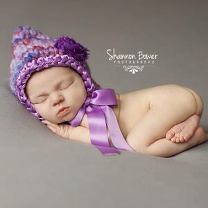 May include: A newborn baby wearing a purple and blue crocheted hat with a matching bow. The baby is curled up on a grey surface, with eyes closed. The hat has a flower on top.
