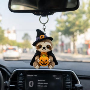 May include: A crocheted sloth hanging ornament featuring a brown and cream sloth wearing a black witch hat with a purple band and a black cape. The sloth holds a small orange jack-o'-lantern with a black face, attached to a silver metal keyring.