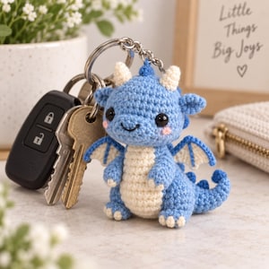 May include: A small, handmade crochet dragon keyring featuring a light blue body, a cream-coloured belly, and cream-coloured horns and wing accents. The dragon has black safety eyes, pink stitched cheeks, and is attached to a silver metal keyring.