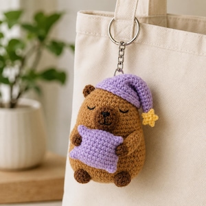 May include: A brown crocheted capybara keychain featuring a purple sleeping cap with a small yellow star at the tip. The capybara has closed eyes and is holding a small purple crocheted pillow, attached to a silver metal keyring and chain.