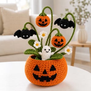 Pode incluir: Um arranjo de crochê para o Halloween num vaso grande em forma de abóbora laranja. O conjunto apresenta duas abóboras laranja mais pequenas, dois morcegos pretos, um fantasma branco e várias margaridas brancas com centros amarelos.
