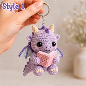 May include: A purple crocheted dragon keychain featuring a small pink book held in its hands, light blue wing accents, cream-colored horns and claws, and black safety eyes. The text Style 1 is visible in the upper left corner of the image.