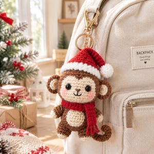 Pode incluir: Um porta-chaves de macaco em croché com corpo castanho e bege, cachecol vermelho e gorro de Pai Natal vermelho com pompom branco. O macaco tem olhos pretos, bochechas rosadas e cauda enrolada. Está preso a um mosquetão e argola dourados.