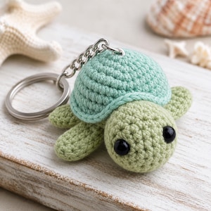 May include: A crochet turtle keyring featuring a light mint green shell and a pale olive green body. The turtle has two large, round, glossy black eyes and is attached to a silver-toned metal split ring by a short silver chain link connector.