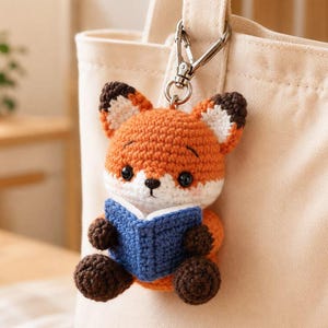 May include: A crochet fox keyring featuring an orange and white fox with dark brown ears and paws, holding a small blue book. The fox has black bead eyes and is attached to a silver-toned metal lobster clasp, hanging from a light beige fabric bag.