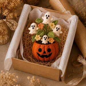 May include: A crocheted pumpkin pot with a jack-o'-lantern face containing four small white ghosts, green leaves, and small orange and yellow flowers. The item is nestled inside a cardboard gift box lined with white tissue paper and wood shavings.