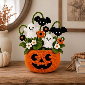 Può includere: Un vaso a forma di zucca arancione all'uncinetto con un volto nero da jack-o'-lantern, riempito con foglie verdi, margherite bianche, fiori arancioni, due fantasmi bianchi con occhi neri e sorrisi, e due pipistrelli neri con occhi bianchi.