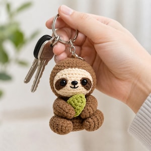 May include: A crocheted sloth keychain featuring a brown body, a cream-colored face with dark brown eye patches, and black plastic eyes. The sloth is holding a small, textured light green leaf and is attached to a silver metal keyring with two keys.