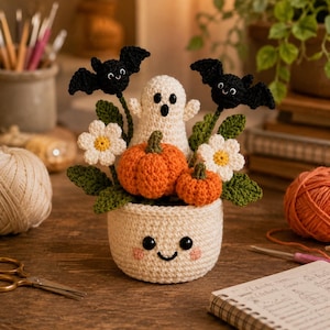Puede incluir: Un arreglo de Halloween tejido a ganchillo en una maceta color crema con una cara sonriente. El conjunto incluye un fantasma blanco, dos calabazas naranjas, dos margaritas blancas con centro amarillo y dos murciélagos negros de lana.