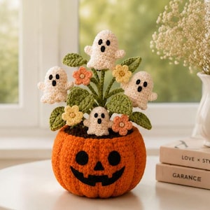 May include: A crocheted orange pumpkin pot with a black jack-o'-lantern face, containing four small white crocheted ghosts, green leaves, and small yellow and peach-colored flowers. The arrangement is crafted from yarn with visible stitches.