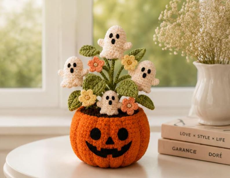 May include: A crocheted orange pumpkin pot with a black jack-o'-lantern face, containing four small white crocheted ghosts, green leaves, and small yellow and peach-colored flowers. The arrangement is crafted from yarn with visible stitches.