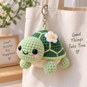 Puede incluir: Un llavero de tortuga de ganchillo con cabeza verde claro, mejillas rosadas, ojos negros y un caparazón verde oscuro con una flor blanca y amarilla. La tortuga está unida a una anilla de metal plateado y cuelga de un bolso de lona color crema.