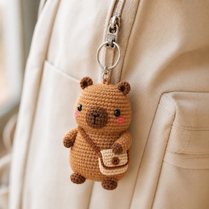 May include: A brown crocheted capybara keychain featuring a small beige crossbody bag, black bead eyes, and pink embroidered cheeks. The figure is attached to a silver metal split ring and is hanging from the zipper of a beige backpack.