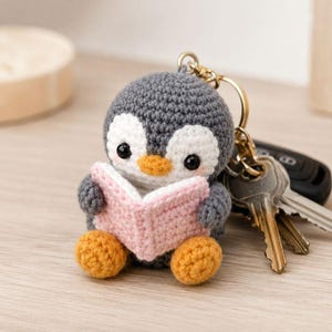 Può includere: Un portachiavi all'uncinetto a forma di piccolo pinguino grigio e bianco che tiene in mano un libro aperto rosa chiaro. Il pinguino ha becco e piedi arancioni vivaci, occhi a perla neri e guance rosa. È fissato a un anello portachiavi dorato.
