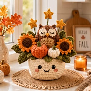 May include: A crocheted arrangement featuring a brown owl with closed eyes, two sunflowers, an orange pumpkin, a white pumpkin, and three yellow stars, all contained within a cream-colored crocheted pot with a smiling face and pink cheeks.