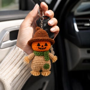 May include: A crochet scarecrow keychain featuring a round orange pumpkin head with a black stitched jack-o'-lantern face, a brown hat, a green scarf, and a tan body with a green patch on the leg and frayed yellow yarn at the hands and feet.