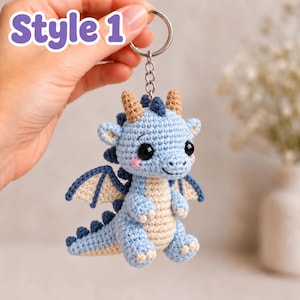 May include: A light blue and beige crochet dragon keyring featuring large black eyes, small horns, and wings. The dragon has a textured yarn surface with a cream-coloured belly and a metal ring attachment. The text "Style 1" is visible in the upper left corner.
