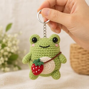 May include: A green crocheted frog keychain featuring a round cream-colored belly, black plastic eyes, pink stitched cheeks, and a small red strawberry-shaped bag with a brown strap worn across the body, attached to a silver metal ring.