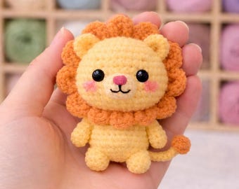 May include: A handmade crochet lion with a yellow body and an orange mane. The lion has black eyes, pink cheeks, and a pink nose. The lion is a small, soft toy, perfect for a child's room or as a decorative item.