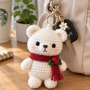 May include: A cream-colored crochet bear keychain featuring a dark red scarf with green holly leaves and red berries. The bear has black eyes, a brown embroidered nose, and pink cheeks. It is attached to a gold-toned metal ring with a white flower charm.
