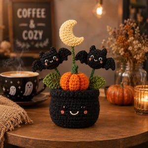 May include: A crocheted black pot with a smiling face containing an orange pumpkin, two black bats with smiling faces, and a yellow crescent moon on green stems. The pot features black yarn, pink stitched cheeks, and black plastic eyes.