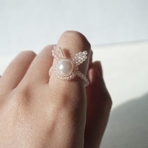 Puede incluir: Un delicado anillo hecho a mano con una perla central rodeada de pequeñas cuentas transparentes. El diseño del anillo incluye detalles de cuentas que se asemejan a orejas de conejo. El anillo se lleva en un dedo, mostrando sus intrincados detalles.